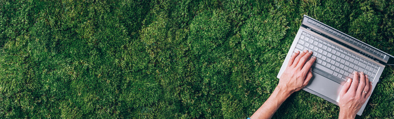 Hands on computer, laptop keyboard over green grass, moss forest background. Top view. Mindfulness, biophilic design, unplug concept. Digital detox. Summer office, work on vacation, freelance concept