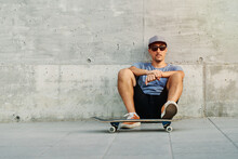 Confident Male Skater In Stylish Outfit Sitting Near Urban Building With Skateboard And Looking At Camera