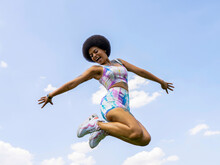 Low Angle Of Delighted African American Female In Summer Outfit In Moment Of Jumping Above Ground On Background Of Blue Sky