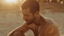 Side View Of Serene Handsome Male With Naked Torso Sitting On Sand On Shore And Enjoying Summer At Sunset