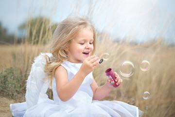 Portrait of a beautiful angel girl with wings in white dress. She is joyfully  blowing soap bubbles. 