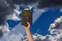 Arm Raised Into The Air With Hand Reaching Up And Holding The Holy Bible. Dark Blue Sky And A Ray Of Light Coming Through Dramatic Blurred Clouds . Symbolic Posture Of Prayer, Devotion And Worship.