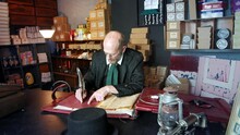 Ebeneezer Scrooge In His Counting House Shop. The Man Is In Victorian Clothing From Charles Dickens A Christmas Carol. Using A Quill To Write In A Leather Bound Book.