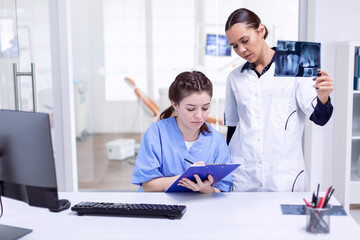 Dentist nurse taking notes about teeth hygine while doctor is holding x-ray. Stomatolog and her assistant in reception of dental clinic looking at teeth radiography.