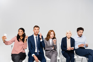 Wall Mural - Multicultural businesspeople talking on smartphones and taking selfie on chairs isolated on grey