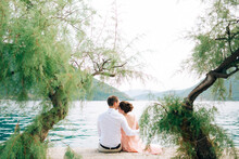 Bride And Groom Are Sitting Among Trees And Hugging On The Pier In Perast