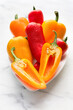Brightly coloured close up of seven red and yellow condensation-covered baby peppers arranged in a small white dish with the front one cut in half against a bright white marble background.