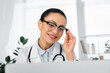 © LIGHTFIELD STUDIOS - Smiling african american doctor touching eyeglasses while looking at laptop on blurred foreground