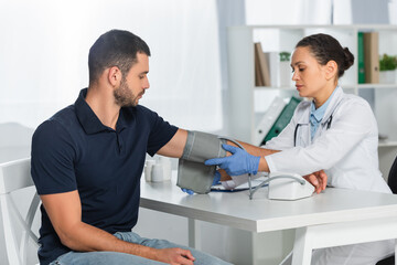 Wall Mural - African american doctor putting tonometer on hand of patient, while sitting at workplace on blurred background