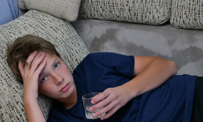 Cute boy in a blue t-shirt lying on the sofa and holding a glass of water in his hand.Portrait of a boy with a glass of water holding his head with his hand.