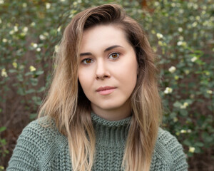 Young beautiful 27 years old woman in a sweater, standing outdoors against a background of greenery, sincerely and expressively looking into the camera. Close-up photo