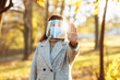 © Konstantin Zibert - Young woman wearing a face shield to protect from coronavirus desease stands in the park during autumn season on a sunset. Female outdoors shows no sign with hand. Quarantine pandemic fall concept.