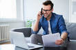 © Bartek Szewczyk/Westend61 - Smiling man with documents and laptop on sofa talking on cell phone
