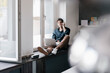 © Kniel Synnatzschke/Westend61 - Smiling young man on the phone sitting with laptop on window sill in a loft