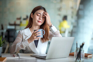 Charming asian female office worker working on a laptop computer and enjoying drinking coffee in a modern office.