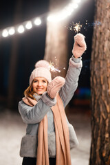 Wall Mural - Christmas sparkles in hands. Cheerful young woman celebrating holding sparkles in the winter forest. Festive garland lights. Christmas, new year.	