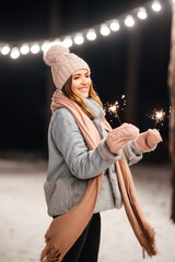 Wall Mural - Cheerful young woman celebrating holding sparkles in the winter forest. Festive garland lights. Christmas, new year.