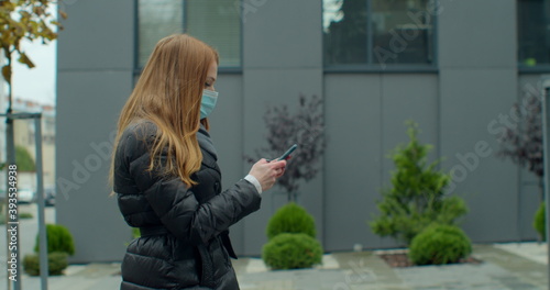 Fotografía Young redhead woman in protective medical mask walks down to the street uses phone texts scrolls surfs the internet