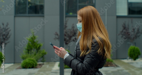 Cuadros en Lienzo Young redhead woman in protective medical mask walks down to the street uses phone texts scrolls surfs the internet