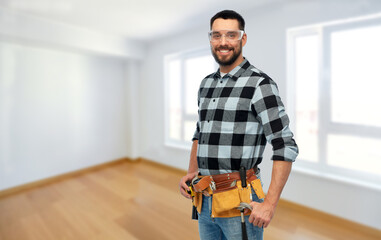 repair, construction and building concept - happy smiling male worker or builder in goggles with tool belt over room at new home on background