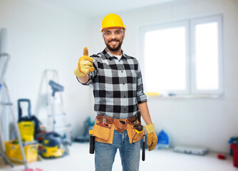 Wall Mural - profession, construction and repair concept - happy smiling male worker or builder in helmet and goggles showing thumbs up over room with building equipment background