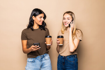 Portrait of two smiling women using cellphones while drinking coffee takeaway isolated over beige background