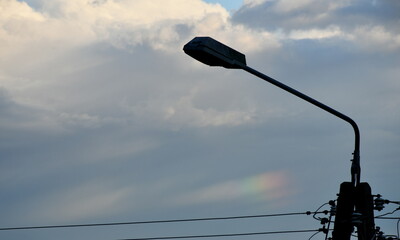 A close up on an old metal lamp post with a concrete base and a bulb  cover in front of it with a cloudy stormy sky behind the item and a slightly visible rainbow between the clouds seen in Poland