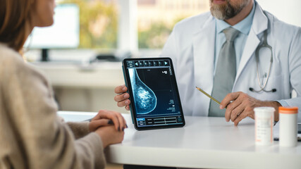 Wall Mural - In a Doctor's Consultation Room: Friendly Professional Doctor Uses Digital Tablet Computer to Show Breast Scan to a Female Patient and Explains Importance of Breast Cancer Prevention. Focus on Tablet