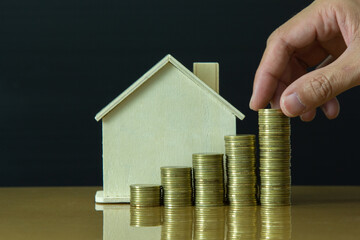 male hand putting golden coin stack growth up with wooden house in background, saving concept