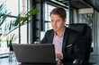 © VideoFlow - Young businessman wearing black suit, sitting to table with laptop. Office manager working in modern office with glass windows blurred background, glass of water on the table, concept of work