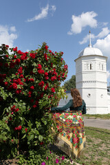 Wall Mural - Blossoming bush: roses in summer garden. Red flowers. Russian girl in traditional national folk Pavlovo Posad shawl. Woman in Suzdal town, Vladimir region, Russia. Russian style in fashion. Red roses