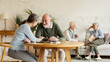 © Comeback Images - Elderly man and woman sitting at table and enjoying joyful talk, another senior couple communicating in background sitting on sofa in common room of assisted living home