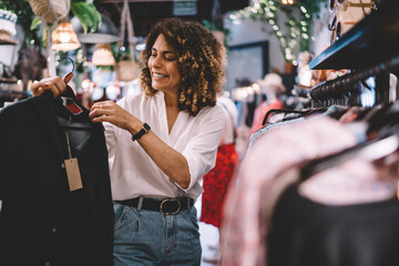 Poster - Cheerful female standing near hangers with brand clothes and laughing during Black Friday shopping with discounts, happy hipster girl enjoying pastime for update her wardrobe spending day in boutique