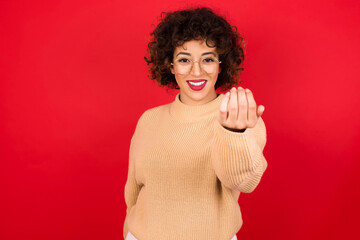 Young beautiful Arab woman wearing beige sweater against red background, inviting you to come, confident and smiling making a gesture with hand, being positive and friendly.