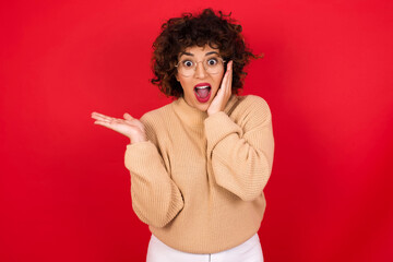 Sticker - Positive glad Young beautiful Arab woman wearing beige sweater against red background says: wow how exciting it is, indicates something.  One hand on his head and pointing with other hand.