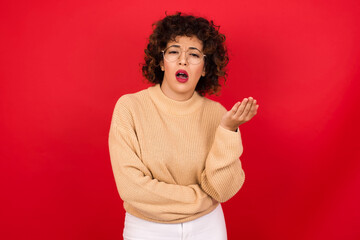 Wall Mural - Studio shot of frustrated Young beautiful Arab woman wearing beige sweater against red background gesturing with raised palm, frowning, being displeased and confused with dumb question.