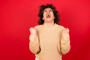 Wall Mural - Young beautiful Arab woman wearing beige sweater against red background looks with excitement up, keeps hands raised, notices something unexpected.