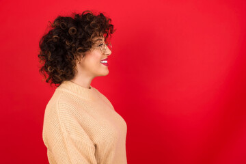 Wall Mural - Profile of smiling Young beautiful Arab woman wearing beige sweater against red background with healthy skin, has contemplative expression, ready to have outdoor walk.