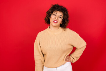 Wall Mural - Studio shot of cheerful Young beautiful Arab woman wearing beige sweater against red background keeps hand on hip, smiles broadly.