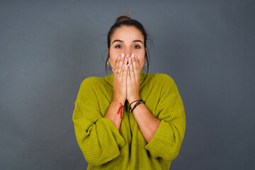 Wall Mural - Vivacious Young beautiful hispanic woman wearing green sweater against gray background, giggles joyfully, covers mouth, has fun alone,  has natural laughter, hears positive story or funny anecdote