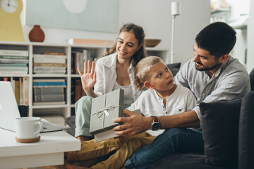 Canvas Print - young happy family with one kid sitting on sofa and opening gifts while having video call on computer