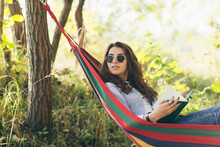 Young Beautiful Girl Resting In A Colored Hammock In The Park And Reading A Book