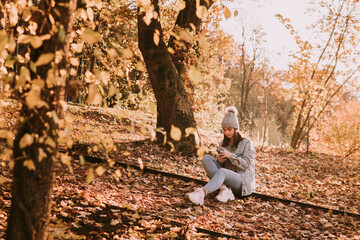 A modern young teenage brunette girl with a hat, dressed in jeans sits in a park on the ground with her legs crossed and uses a smartphone. Autumn walk in the woods