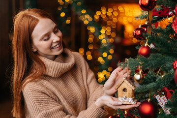 Portrait of smiling young woman decorating Christmas tree beautiful toy at cozy living room. Cheerful red-haired lady putting xmas ornament on new year tree. Preparation home to holiday celebration.