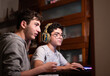 © Massimo Parisi - Staying at home: Two Caucasian brothers keep busy with their favorite video game. One is sitting with the gaming headset on his head and the other is standing next to him to suggest moves to take.