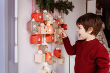 Happy Excited Little Boy Waiting To Start Opening Handmade Advent Calendar Hanging On Wall. Sustainable Christmas, Upcycling, Zero Waste, Kids Seasonal Activities