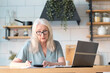 © yavdat - Senior woman using laptop for websurfing in her kitchen. The concept of senior employment, social security. Mature lady sitting at work typing a notebook computer in an home office.