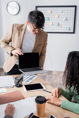 Wall Mural - Hispanic office worker showing laptop with blank screen to female colleague sitting at workplace in office