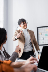 Wall Mural - Cheerful hispanic office worker showing thumb up, standing near workplace with blurred co-workers on foreground