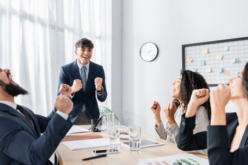 Wall Mural - Excited hispanic executive with yes gesture standing near co-workers sitting at workplace during business meeting